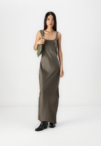 Rochie maxi din satin de culoare verde măsliniu, cu fante laterale, bretele ajustabile și o textură fină. Asortată cu cizme negre cu toc și o geantă verde.