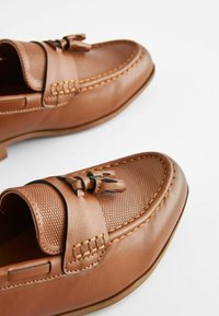 Mocassins en cuir marron avec un panneau texturé, présentant des lacets décoratifs et des coutures contrastées. Fini lisse et design à bout arrondi.
