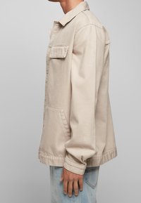 Mężczyzna w beżowej kurtce zapinanej na guziki z flapem na piersiach i bocznymi kieszeniami, w połączeniu z jasnoniebieskimi dżinsami, stojący bokiem.