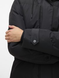 Črna podložena jakna z visokim ovratnikom, teksturirane obdelave, z gumbi na sponki na manšetah in kapuco z vrvico. Roke prekrižane spredaj.