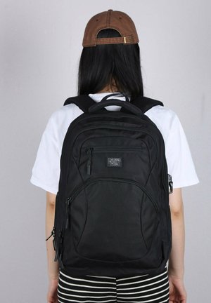 Zaino nero con numerosi scompartimenti con zip, spallacci imbottiti e materiale resistente. Presenta una maniglia superiore e un marchio discreto.