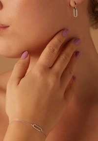 Boucles d'oreilles en argent au design rectangulaire ; peau lisse et claire ; main avec des ongles violets clairs touchant la joue ; bracelet délicat visible.