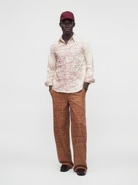 Homme portant une casquette rouge, une chemise crème avec un motif gribouillé rouge, un pantalon marron avec un motif rouge similaire, debout les mains dans les poches.
