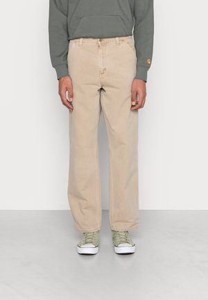 Mann trägt beige gerade geschnittene Hose, grünen Sweatshirt und hellgrüne Turnschuhe, steht vor schlichtem weißem Hintergrund.