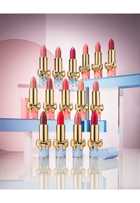 Lippenstifte mit goldenen Tuben und blauen Basen, angeordnet in verschiedenen Pink- und Rottönen, mit Schleifenakzenten und einer reflektierenden Oberfläche.