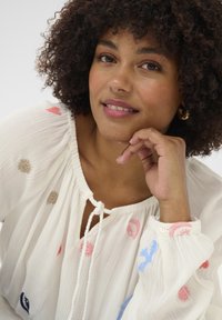 White blouse with a textured fabric, featuring colorful embroidered details in pink, blue, and beige. Ties at the neck and buttoned cuffs.