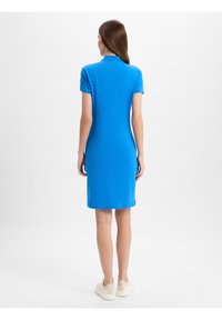 Blaues Polokleid mit kurzen Ärmeln und Kragen, das bis zur Mitte des Oberschenkels reicht. Hergestellt aus glattem Stoff; kombiniert mit weißen Sneakers.