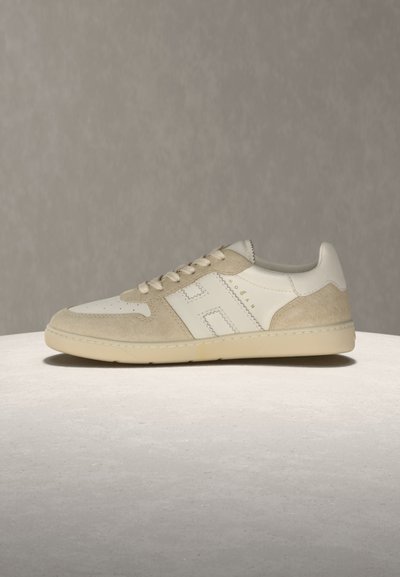 Baskets basses blanches et beiges avec des détails en suède, lacets à l'avant, et logo "H" brodé sur le côté, sur fond neutre.