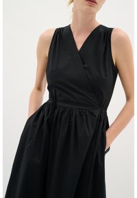 Schwarzes ärmelloses Kleid mit Wickeldesign, elastischem Bund und Taschen. Glatte Textur mit einem fließenden Rock und Schulterfalten.