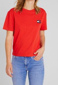 T-shirt in cotone rosso con colletto rotondo, maniche corte e una toppa con logo sul lato sinistro del petto. Abbinato a jeans in denim blu chiaro.