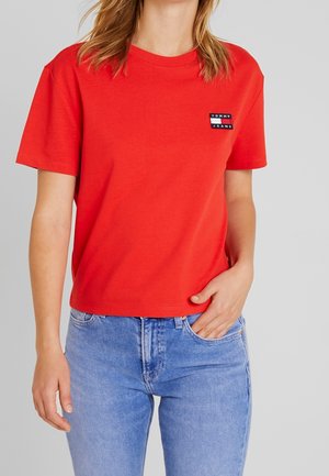 Rød bomulds T-shirt med rund hals, korte ærmer og et logo-patch på venstre bryst. Kombinationen med lyseblå denim jeans.