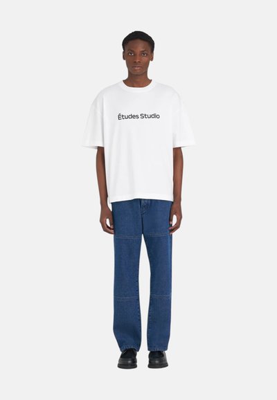 Personne portant un T-shirt blanc surdimensionné avec le texte "Études Studio", un jean bleu ample avec des coutures aux genoux, et des chaussures noires, se tenant devant un fond uni.