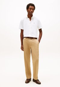 Muž s krátkymi kučeravými vlasmi, oblečený v bielej košeli s krátkym rukávom a gombíkmi, béžových nohaviciach, hnedom opasku a hnedých kožených topánkach, stojaci s rukami v kapsách.