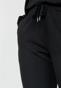 Black joggers made of soft fabric, featuring an elastic waistband with drawstring and silver hardware, side pockets, and a smooth texture.