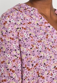 Blouse fleurie en coton violet pastel avec de petites fleurs multicolores, accents noirs, et une texture douce et légère. Col en V.