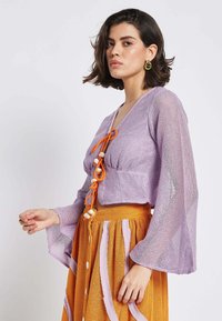 Purple, cropped knit top with bell sleeves and orange ties. Paired with a textured orange skirt featuring pink and white vertical stripes.