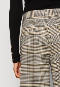 Gros plan d’une personne portant un pantalon à carreaux beige, marron et noir et un haut noir à manches longues avec des boutons dorés sur le poignet.
