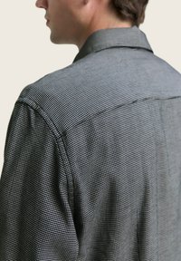 Camisa de cuadros en blanco y negro con cuello puntiagudo, que presenta una tela texturizada y un detalle de costura visible en la costura del hombro.