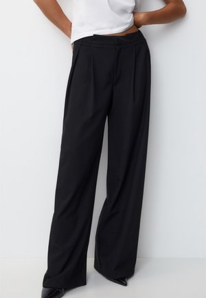 Persoon die losse zwarte wijde broek en puntige zwarte schoenen draagt, staand met handen achter de rug tegen een witte achtergrond.