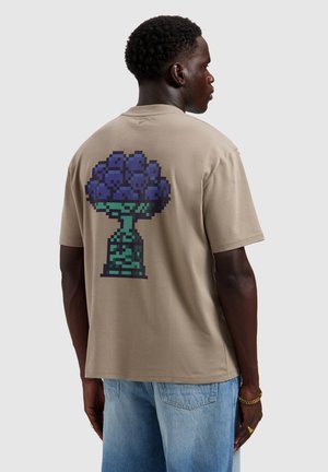 Man wearing beige t-shirt with pixel art trophy holding a cluster of purple grapes on the back, paired with light blue jeans.