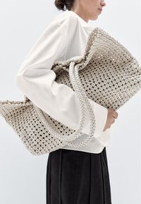 Grote geweven tas in lichtbeige, met een gestructureerd patroon, gevlochten handvaten en een slungelige uitstraling. Gecombineerd met een witte top en zwarte broek.