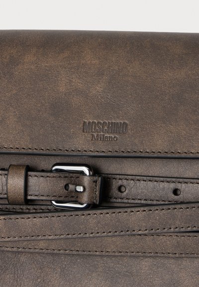 Détail d’un sac en cuir marron avec une boucle argentée et un logo Moschino Milano embossé au-dessus de la sangle cousue.