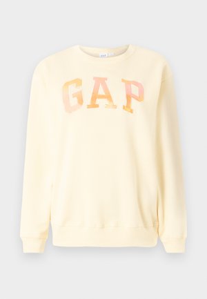 Gul genser laget av mykt stoff, med et overdimensjonert "GAP"-logo i iriserende skrifttype. Rund hals og lange ermer, ribbestrikkede mansjetter.