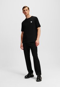 Tricou scurt negru cu mânecă scurtă, featuring un mic logo alb în formă de inimă, asortat cu pantaloni negri și pantofi negri lucioși. Design simplu și minimalist.