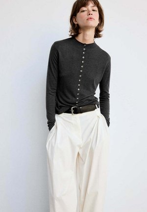 Jeune personne portant un haut sombre à manches longues boutonné, rentré dans un pantalon blanc ample à taille haute avec une ceinture noire, debout contre un mur uni.