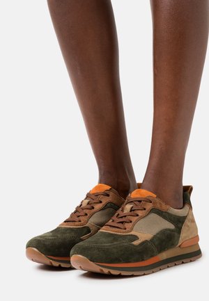 Hnědé a zelené semišové tenisky s oranžovými akcenty, nošené na bosých nohou, zobrazené zepředu na bílém pozadí.