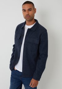 Marineblå flanneljakke med knaplukning foran, to brystlommer og krave. Båret over en hvid t-shirt og sammen med blå jeans.