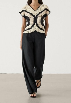 Haut en crochet avec un col en V en crème et noir, présentant des motifs courbes. Associé à un pantalon noir à jambes larges taille haute et des sandales.