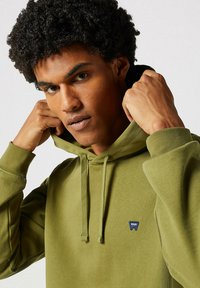 Sudadera verde oliva hecha de una mezcla de algodón, que cuenta con un bolsillo canguro en la parte delantera, capucha con cord�ón y un pequeño parche de logo en el pecho.