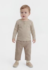 Malček stoji, oblečen v bež črtasto srajco z dolgimi rokavi, svetlo rjave hlače, kremne nogavice in bež natikače na belem ozadju.