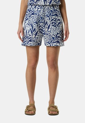Donna che indossa pantaloncini con motivo floreale blu e bianco e sandali marroni con doppie cinghie e fibbie dorate, in piedi davanti a uno sfondo semplice.