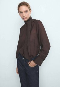 Eine Frau mit blonden Haaren, die zu einem Zopf gebunden sind, trägt eine dunkelbraune Bluse mit langen Ärmeln und einer Schleife am Hals sowie dunkelblaue Jeans. Sie steht vor einem hellen Hintergrund.