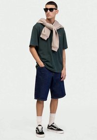 Camiseta de algodón verde oscuro, suéter beige cae alrededor del cuello, pantalones cortos de mezclilla azules, zapatillas negras con suelas blancas y calcetas blancas.