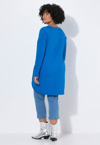 Langärmlige blaue Tunika mit rundem Ausschnitt, kombiniert mit verkürzten Jeans und metallisch silbernen Ankle Boots. Glatte Textur.