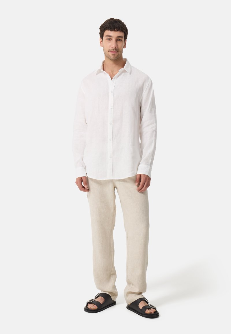 Chemise boutonnée en lin blanc, pantalon en lin beige et sandales noires. Tissu léger et respirant avec une coupe décontractée et des détails de couture visibles.