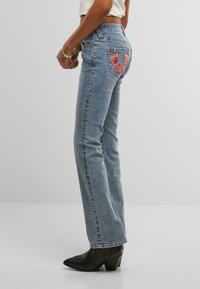 Modri jeans z visokim pasom, ki imajo rožnato vezenino na zadnjem žepu in ravno krojenje.
