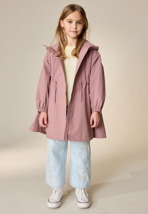 Ragazza giovane in piedi, indossa una giacca con cappuccio malva, una maglietta giallo chiaro, pantaloni ampi azzurri ricamati con motivi floreali e scarpe da ginnastica bianche.