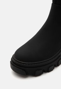 Gros plan frontal d'une botte noire épaisse avec un dessus lisse et une semelle épaisse et robuste sur fond blanc.