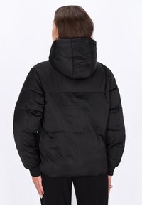Svart puffjacka med huva, med ett quiltat mönster och ribbade ärmslut. Slät, glänsande textur med en avslappnad passform och ingen synlig hårdvara.