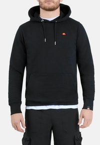 Uomo che indossa una felpa nera con cappuccio, dotata di tasca frontale e piccolo logo arancione-rosso sul petto e sulla manica, abbinata a pantaloni cargo neri.