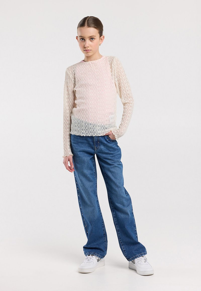 Mädchen steht mit der Hand in der Tasche und trägt ein durchsichtiges langärmliges Spitzenoberteil, blaue Jeans mit seitlichen Knöpfen und weiße Turnschuhe vor weißem Hintergrund.