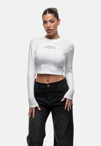 Weißes Langarm-Crop-Top mit geripptem Textur und Details, kombiniert mit hoch taillierten schwarzen Jeanshose mit einzigartigen Nahtmustern.