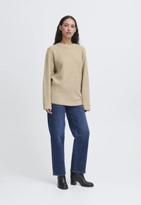 Maglione beige lavorato a maglia con orlo a coste, indossato con jeans blu a gamba dritta e stivaletti neri alla caviglia, in piedi su uno sfondo bianco.