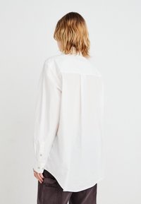 White cotton shirt with long sleeves, buttoned cuffs, and a back yoke. Features a curved hem and relaxed fit. No visible patterns.