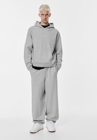 Šedá mikina a zodpovedajúce sweatpants vyrobené z mäkkého materiálu. Mikina má predné vrecko a priestranný strih. Biele tenisky dopĺňajú outfit.