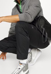 Šedá mikina na zip s zelenými akcenty, černé sportovní kalhoty a šedo-bílé vysoké tenisky. Viditeln ý je černý batoh.